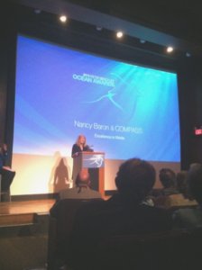 Nancy Baron, with shoulder-length blonde hair, stands behind a podium on stage in front of an audience at the Peter Benchley Awards. The large projection screen behind her has the words "Nancy Baron & COMPASS" written on a blue background. 