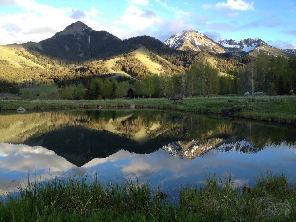 Snow-dusted mountains rise in the distance on a sunny day, with clouds casting the nearest mountain into shadow. Trees and green clearings are visible on the mountainside, while more green trees ring the still lake in the foreground, which is reflecting the mountains and the blue sky.