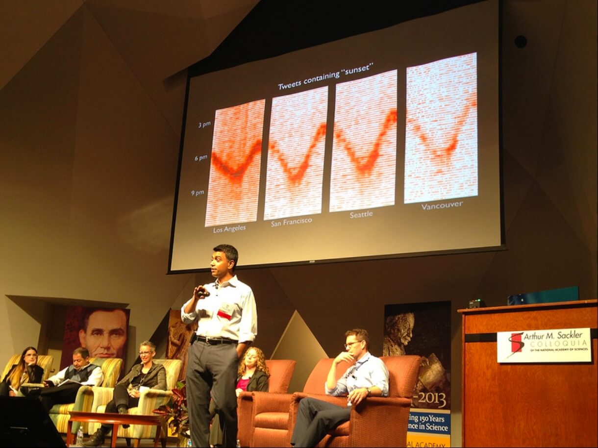 A man with brown skin and short black hair stands in front of a panel of people in armchairs, with a large projector screen above him. He is holding a microphone and addressing the audience, wearing a light-colored collared shirt and gray slacks. The image on the projector is of four graphs lined up in a row; at the top the title says: Tweets containing sunset. Each graph is charting the number of tweets with the word sunset mentioned at different times of day, one graph for each of the cities of Los Angeles, San Francisco, Seattle, and Vancouver. Each graph shows a similar trend and dip in tweets in the middle, though the Los Angeles one is much more pronounced than the Vancouver one.
