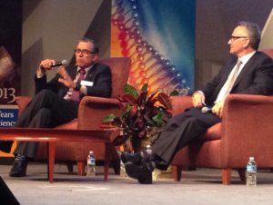 Two older men with light skin, gray hair, and glasses wearing suits sit in armchairs on stage. One is speaking into a microphone while the other listens. 