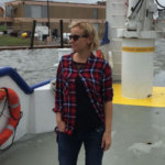 Dr. Natalia Sidorovskaia stands on the deck of a research vessel on the coast, wearing a red plaid shirt and sunglasses. 