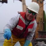 Dr. Tracey Sutton, in a hard hat, foul weather gear, and a life vest, on the deck of a research vessel at sea. 