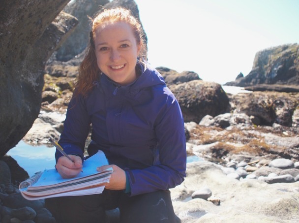 A young graduate student with a blue jacket and long red hair in a ponytail smiles at the camera while taking notes with a pen outside on a rocky beach with seaweed and barnacles.