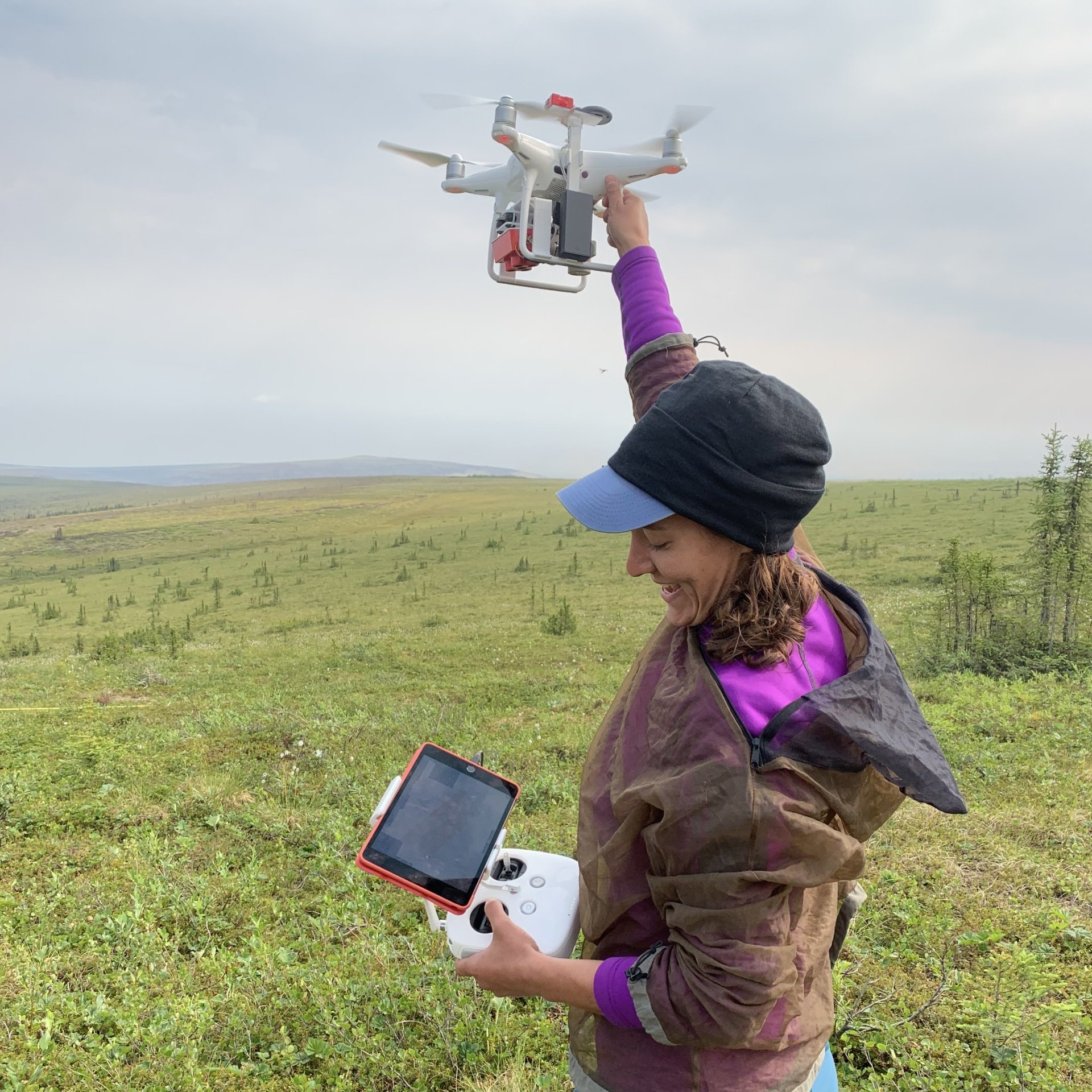 A PhD student stands in a green field, holding a drone overhead to launch it.