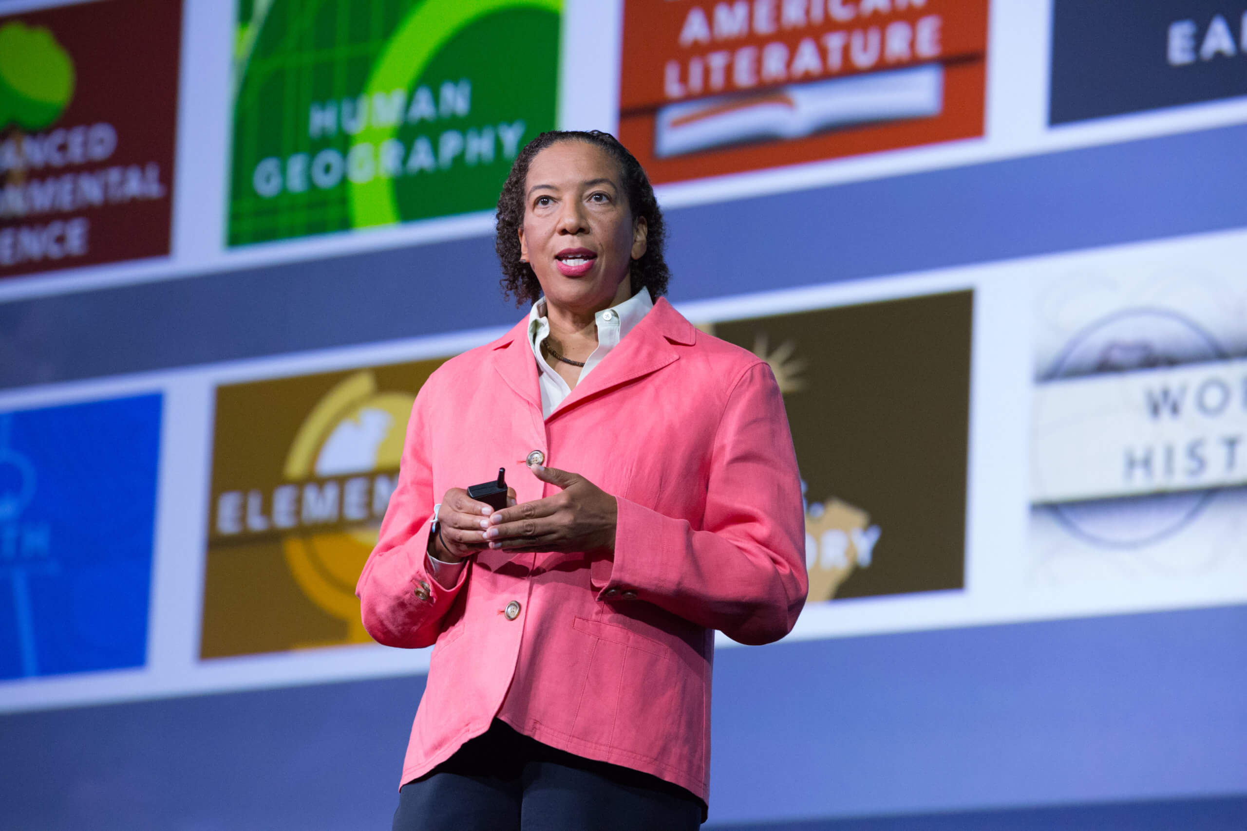 Dr. Dawn Wright presents at an event, with a pink coat, a white shirt, and pink lipstick in front of a screen full of logos.