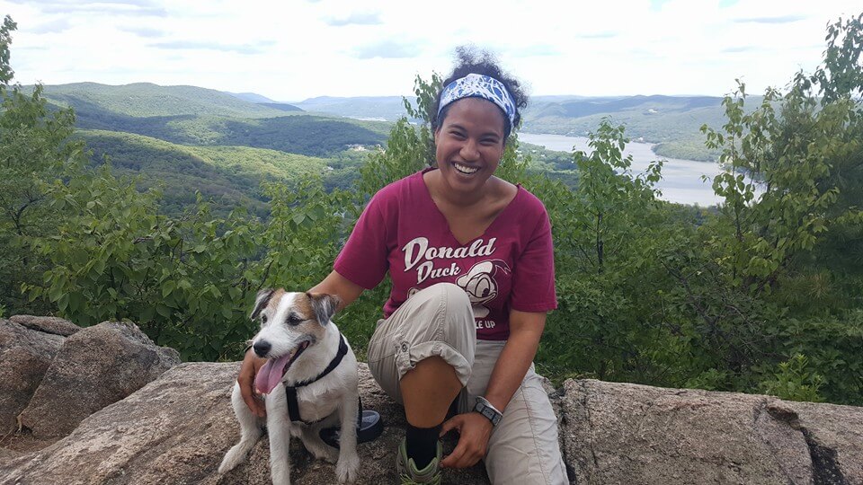 Shanice Bailey is sitting on a rock outside with a Jack Russell Terrier next to her. It looks like she has reached an overlook on a hike, and in the distance we see rolling hills, green trees, and a river going through a gentle valley.