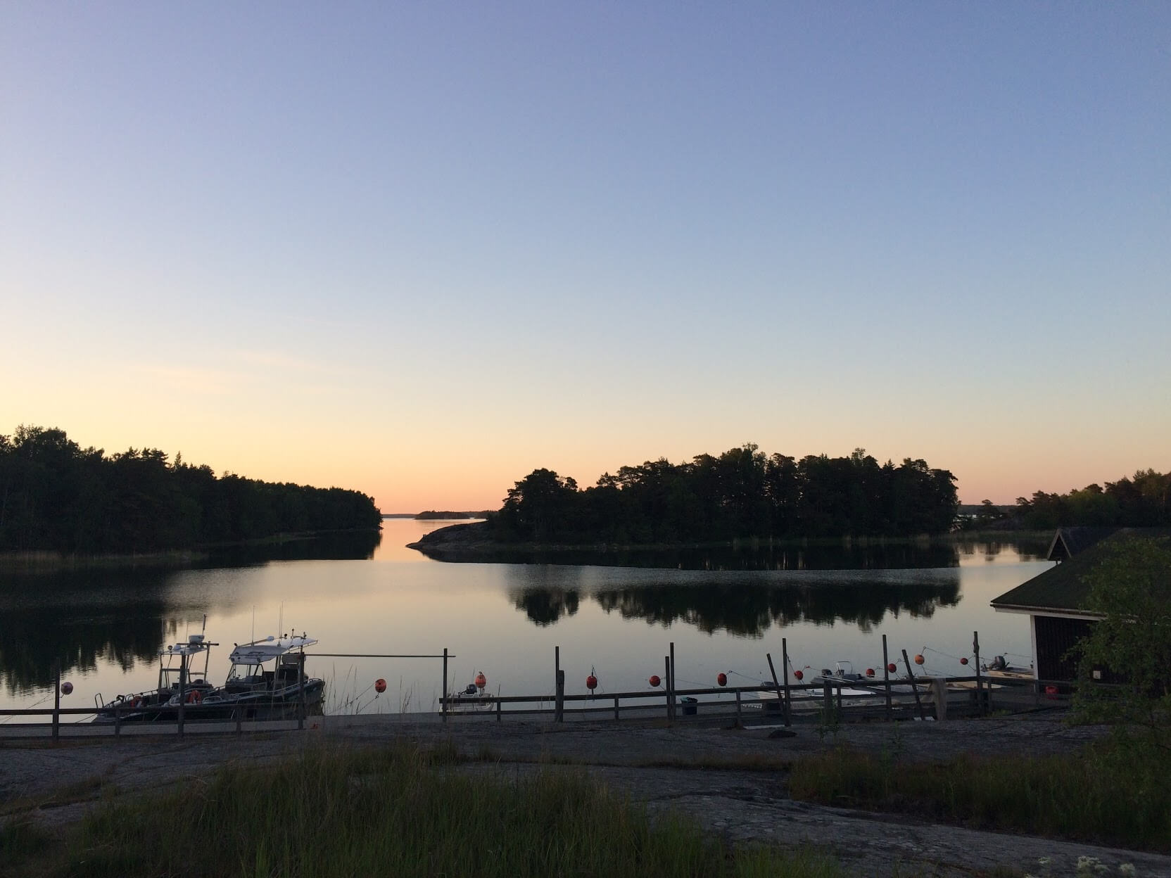 The sun sets behind small, forested islands in the Baltic Sea. A path down to a dock with several motorboats is in the foreground, and the water is still, reflecting the islands and the sky.